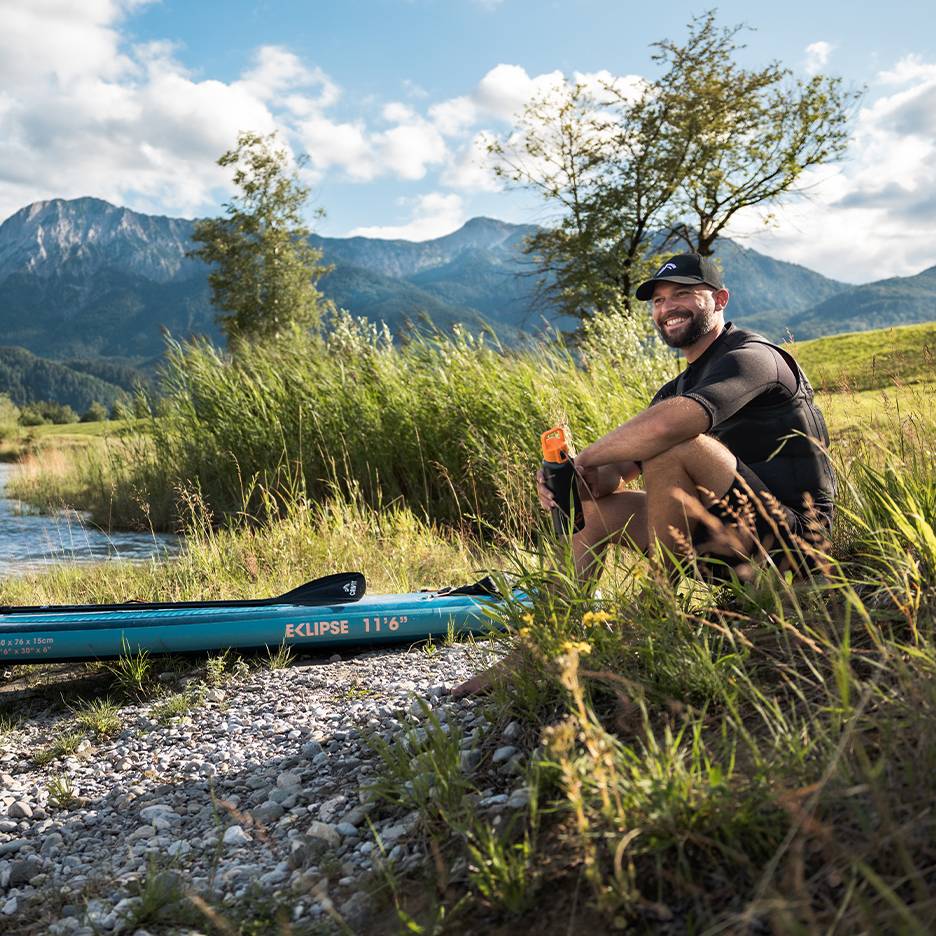 Smiling man in cap and life vest, sitting by a blue paddleboard by the river.