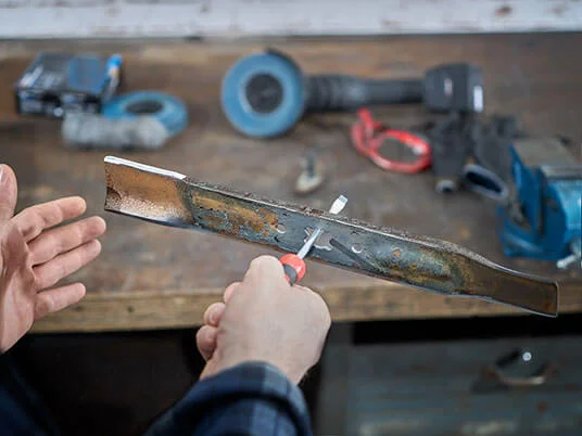Hands sharpening a lawnmower blade on a workbench with tools.