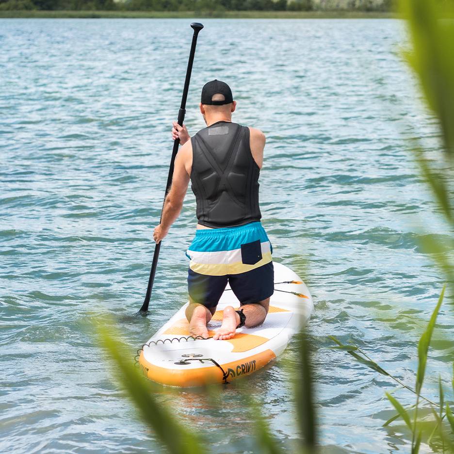 Man paddling on a lake on a Crivit SUP board, wearing a life vest and swim shorts.