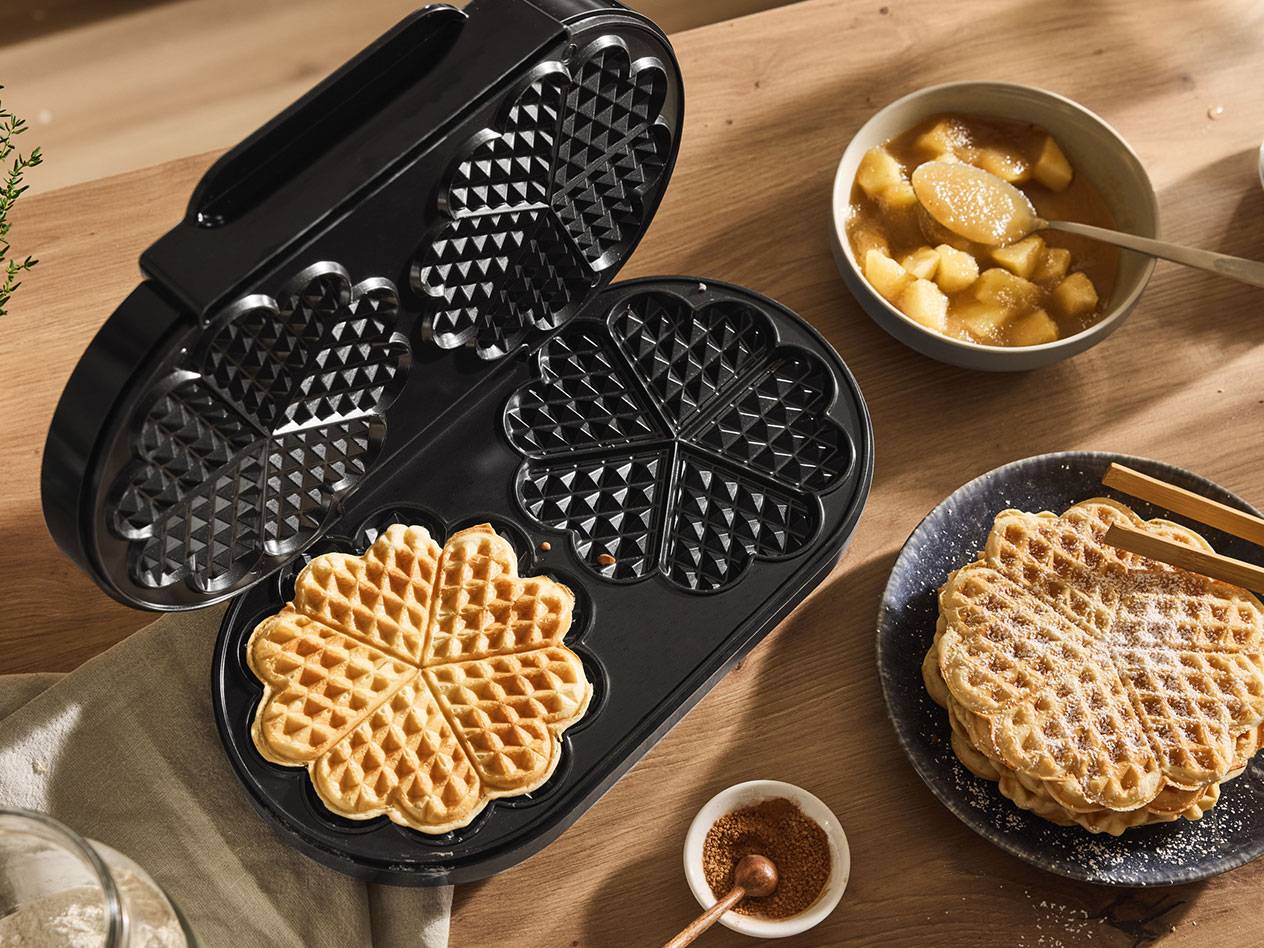 An open waffle maker with a heart-shaped waffle, alongside cooked waffles and apple compote.