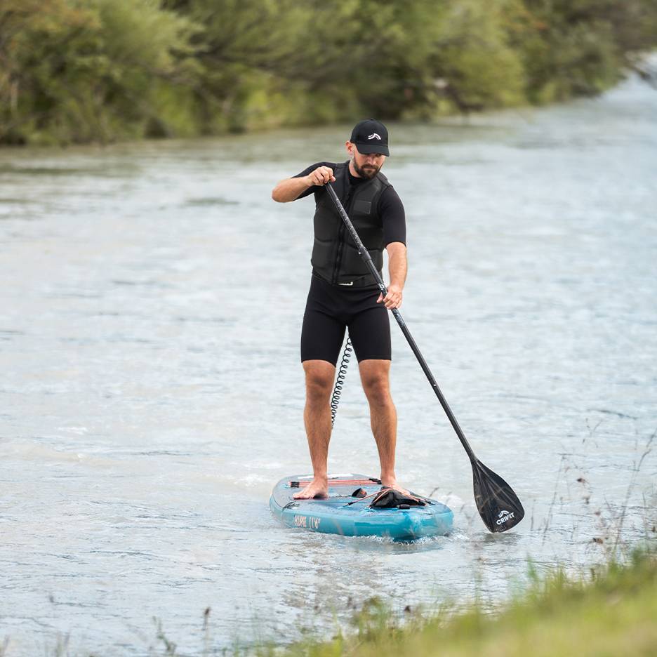 Man paddleboarding on a river, wearing a black wetsuit, life vest, and cap.