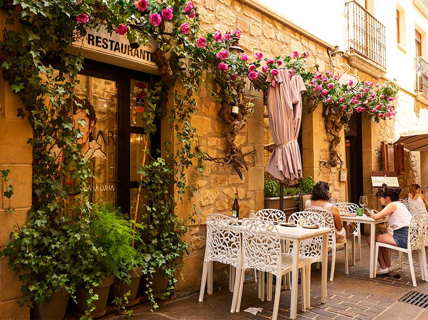 People sitting at tables in front of a restaurant with flowers and climbing plants.