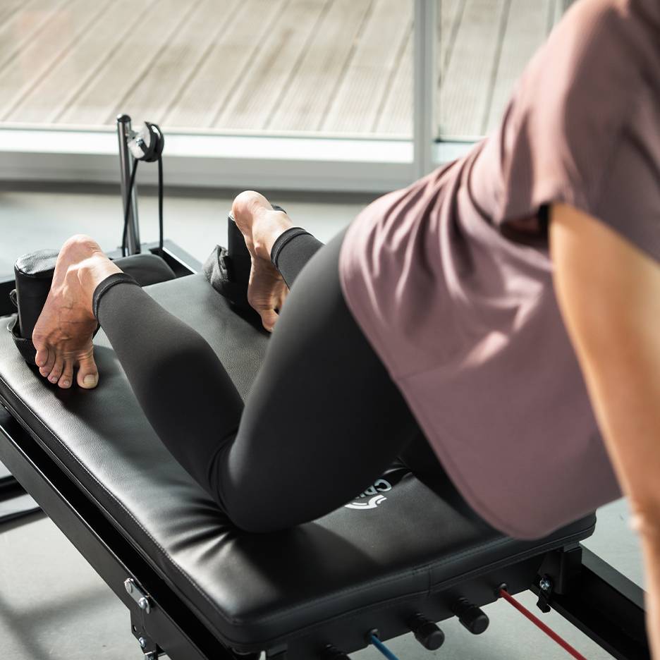 Woman exercising on a black Pilates machine with feet in straps.