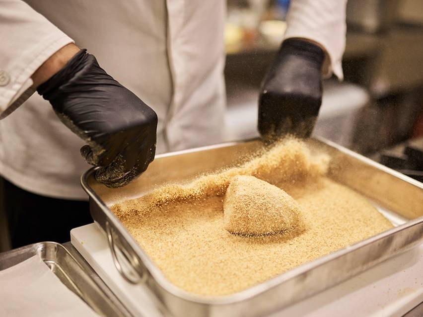 A chef in black gloves breading food in a metal tray.
