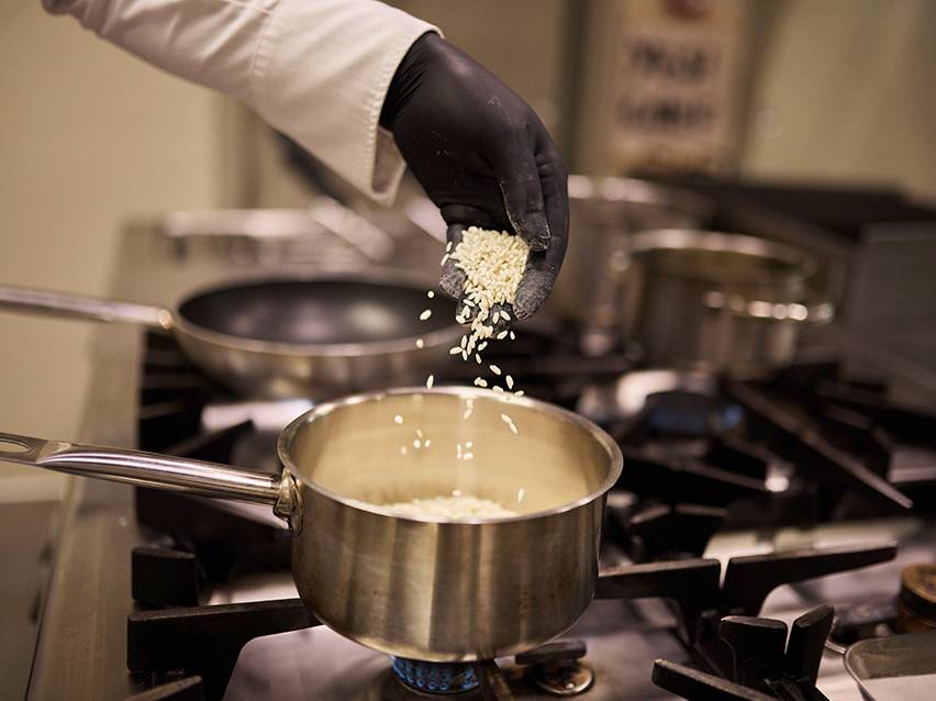 A chef in black gloves pours rice into a pot on a gas stove.