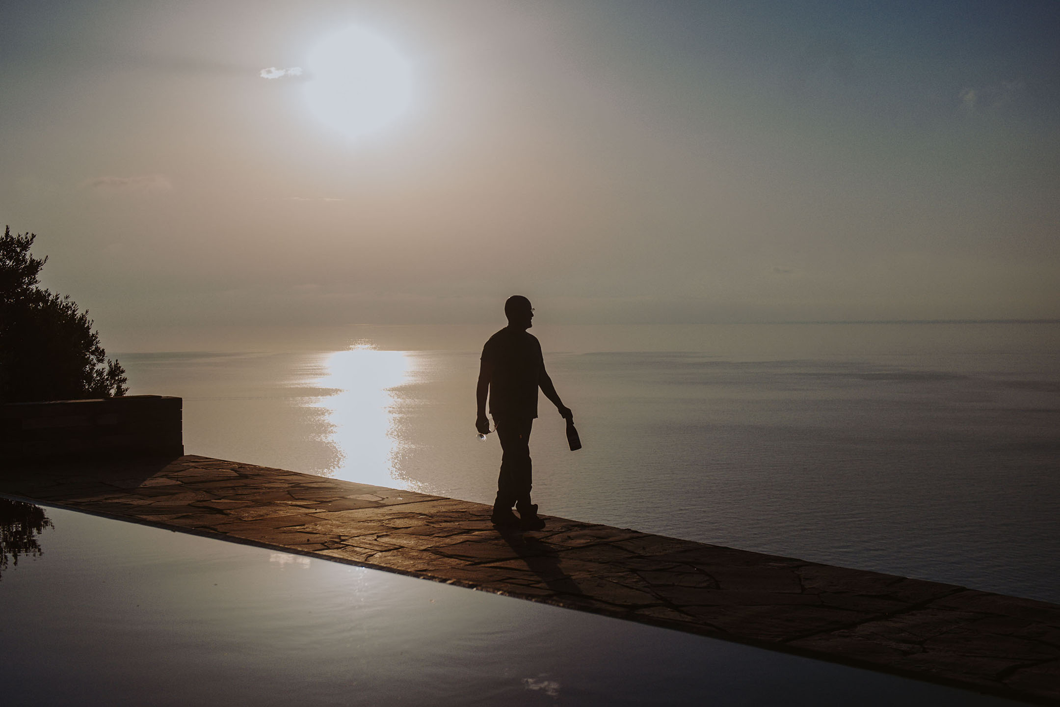 Silhouette of a person holding a bottle and glass by an infinity pool at sunset