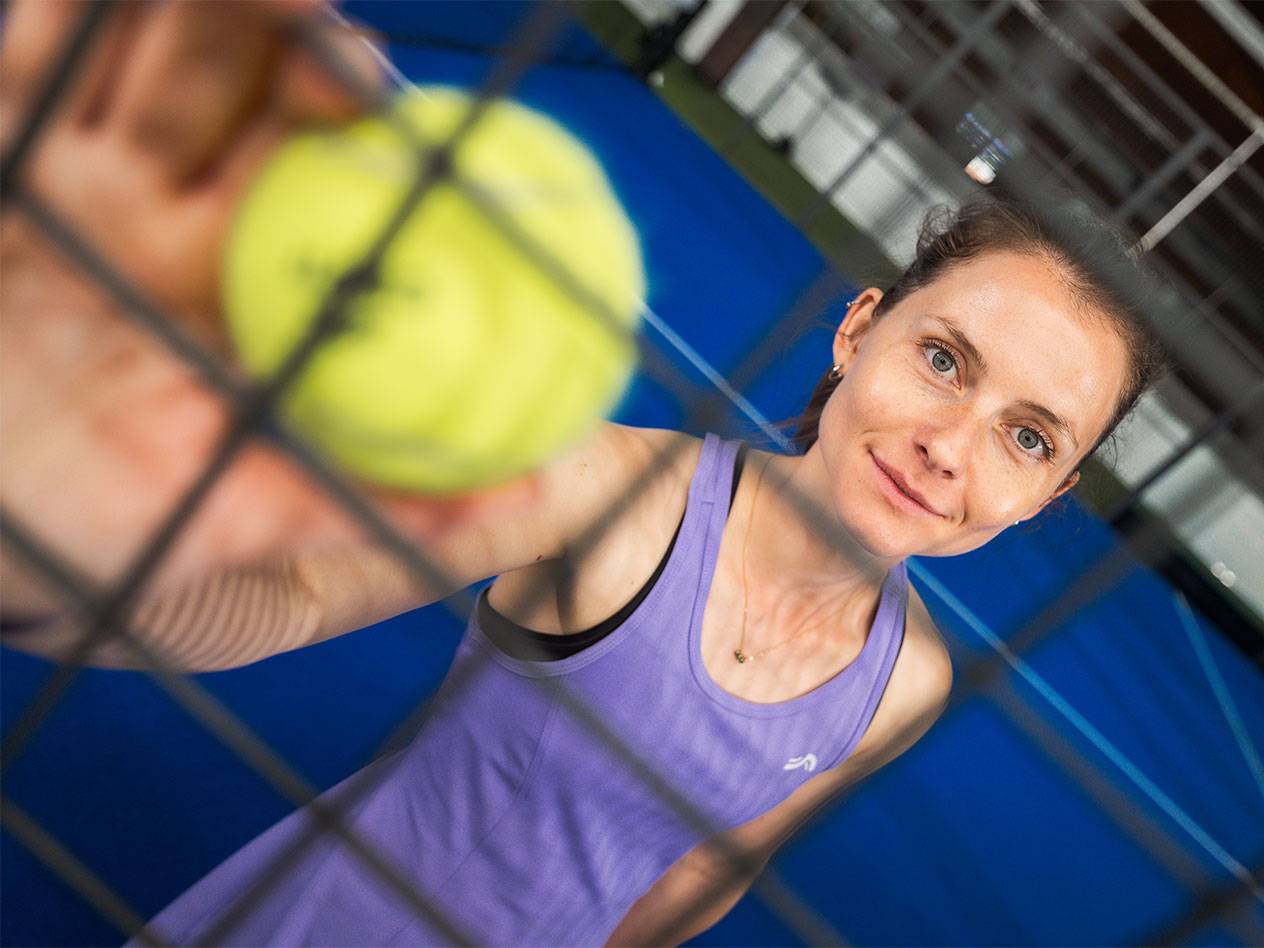 Woman in sportswear holding a padel ball through a net, blue court in the background.