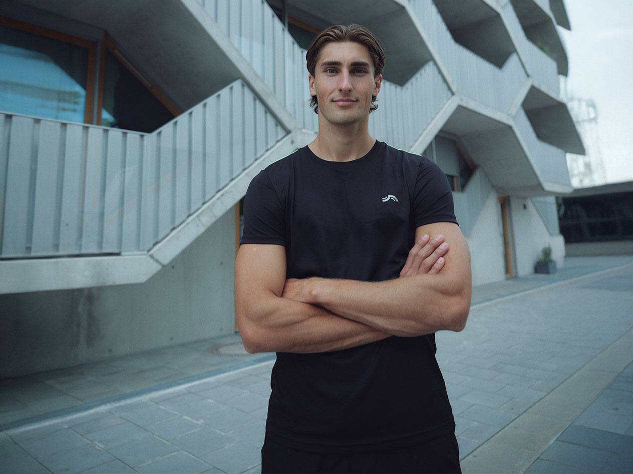 Man in black t-shirt with logo and black shorts, standing outdoors with arms crossed.