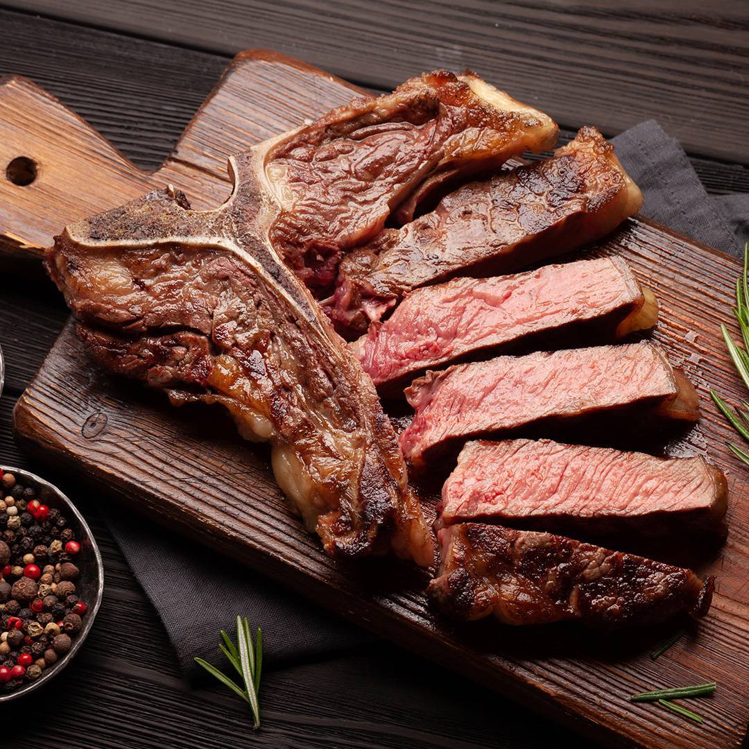 Grilled T-bone steak, sliced, with rosemary and peppercorns on a wooden board.