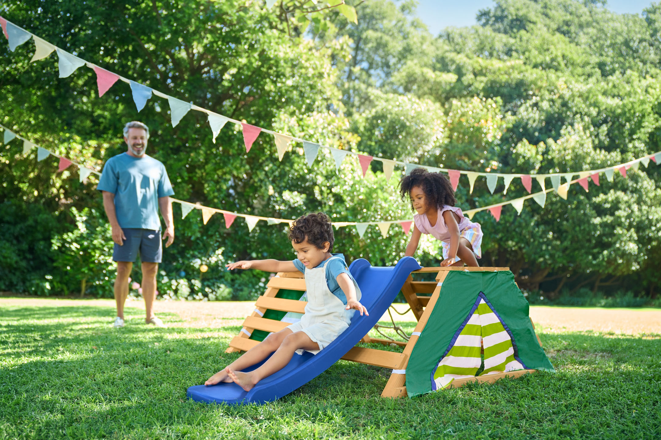 Children playing on a wooden playground with a slide in the garden, with their father in the background.