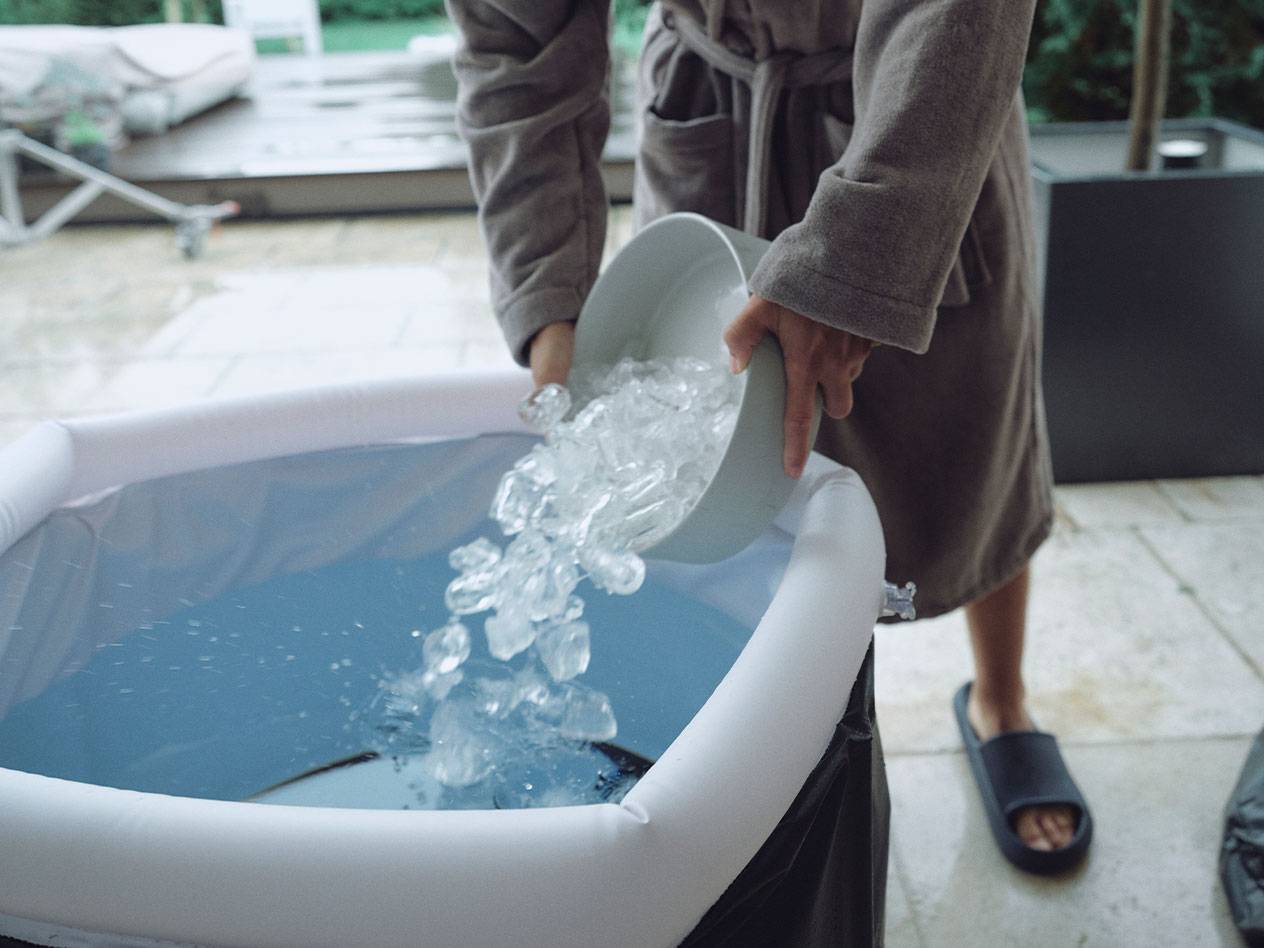 Person in a bathrobe pouring ice into an inflatable outdoor ice bath.