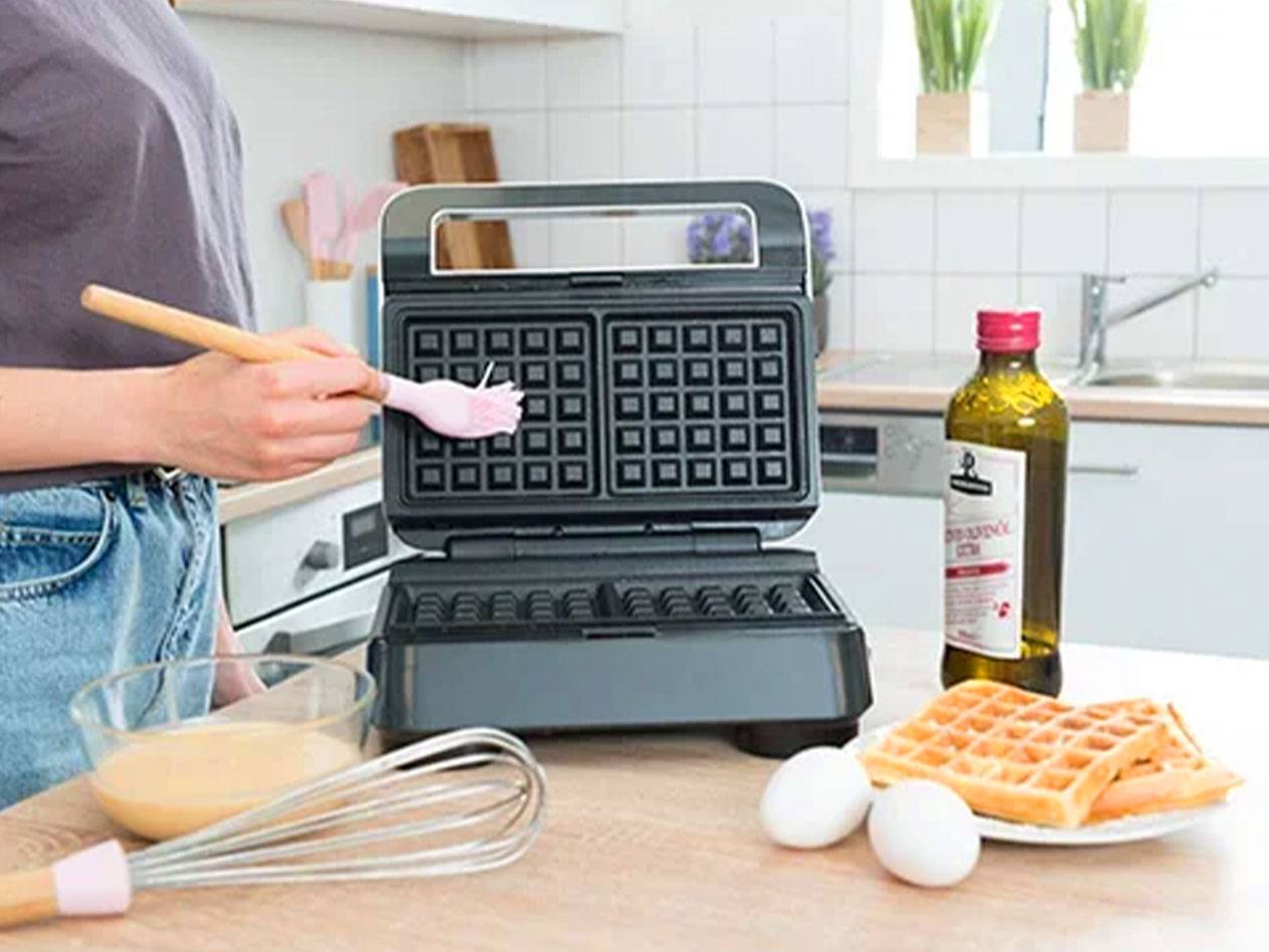 A person brushes a waffle maker with a brush, with ingredients and ready waffles on the table.