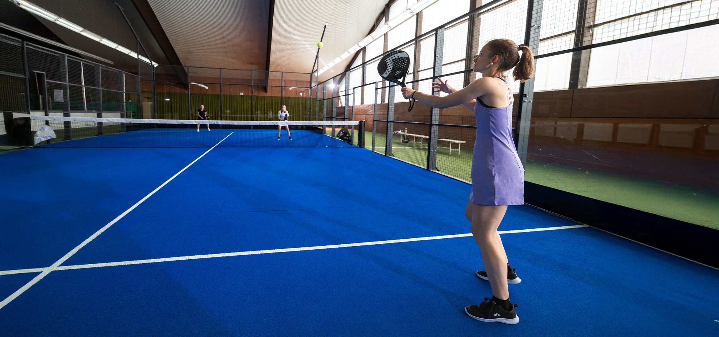 Woman playing padel with Crivit racket, yellow ball, and other players on the court.