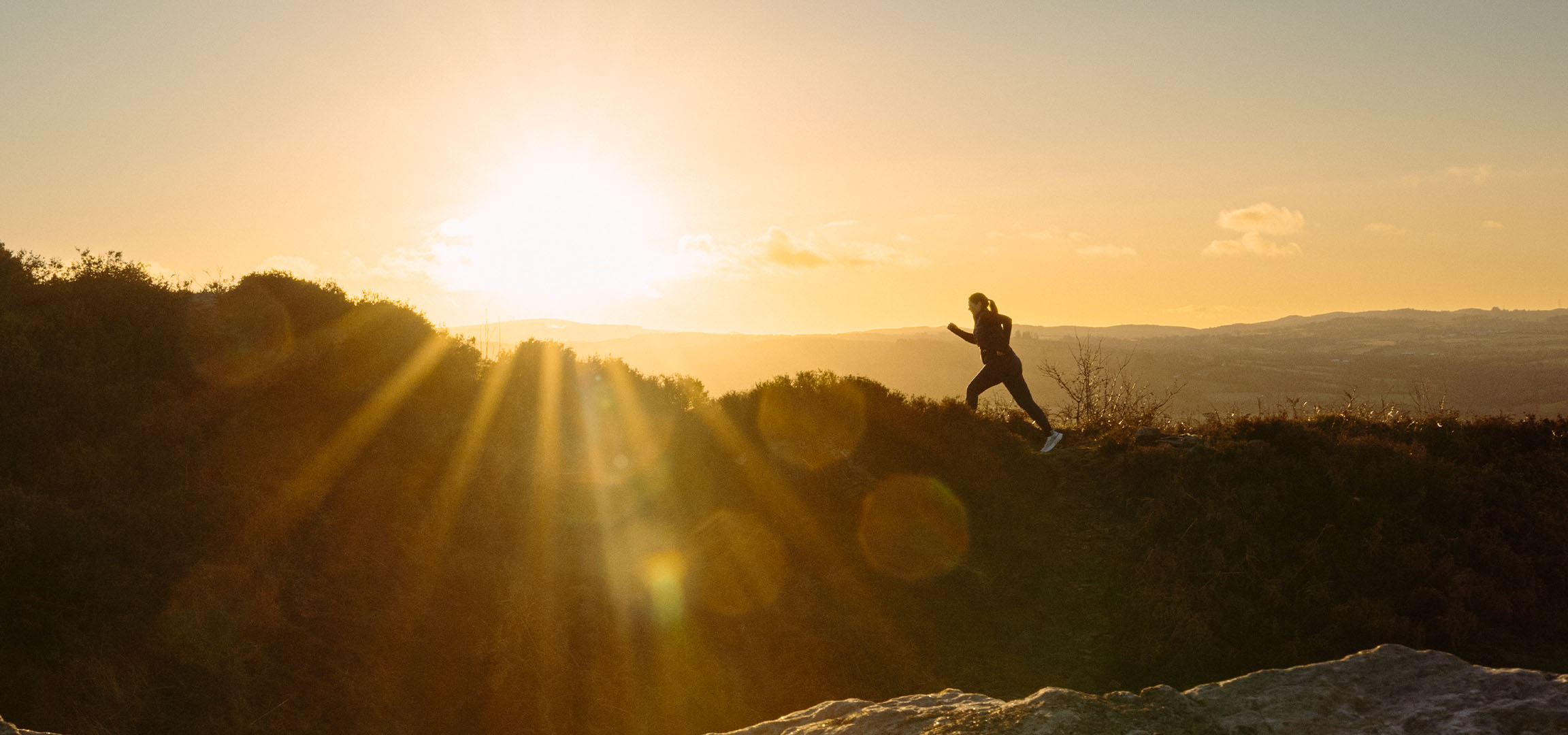 Woman running on a hill at sunset, with sun rays shining through the landscape.
