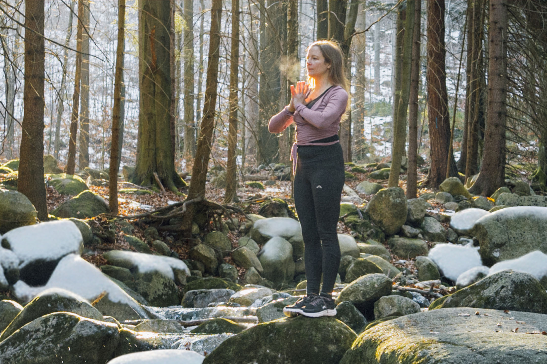Woman in activewear doing yoga in a forest with snow-dusted rocks and trees.