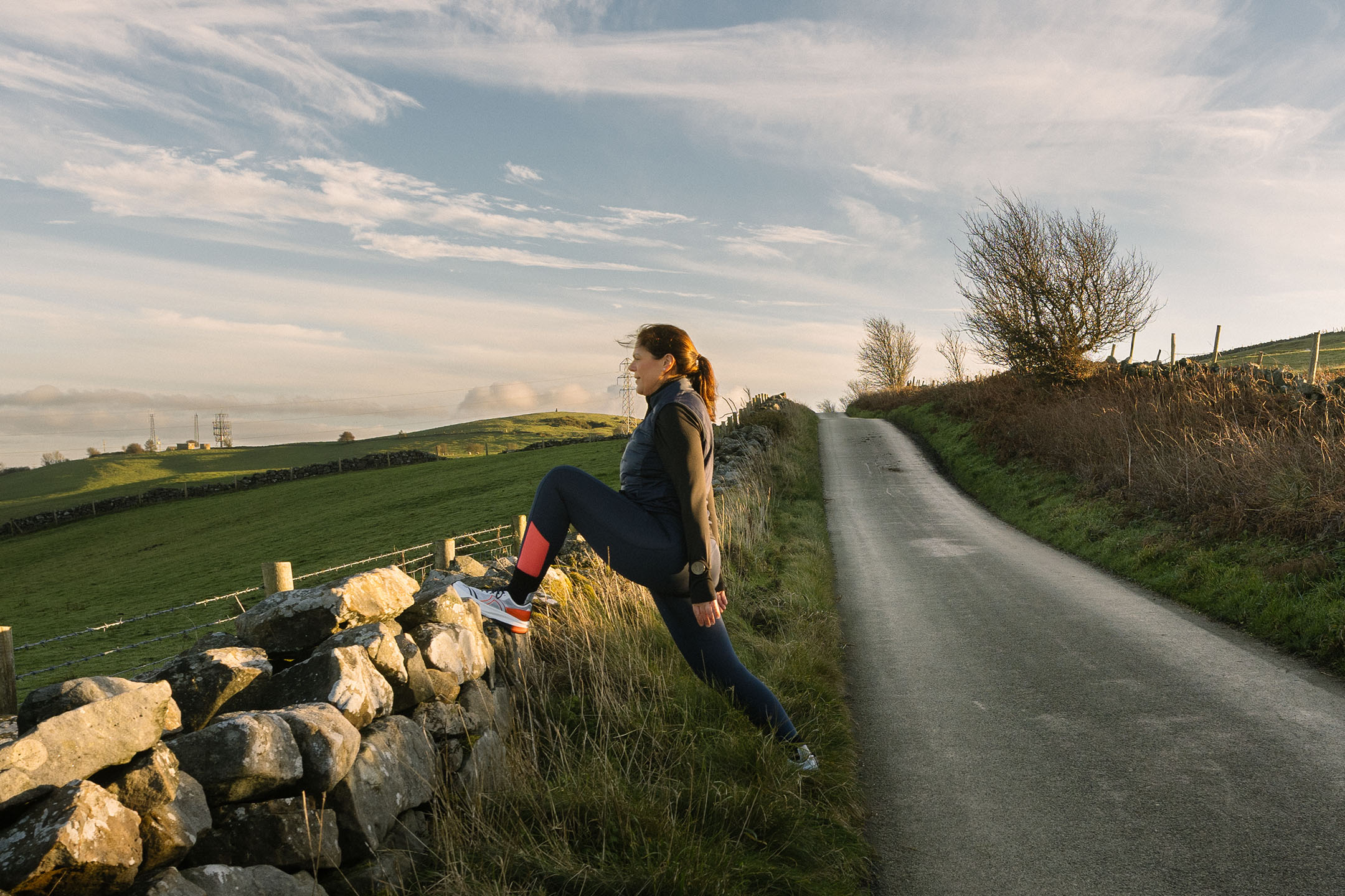Woman in sportswear stretching on a stone wall by a country road.