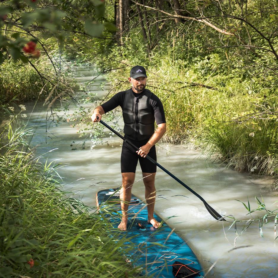 Man in wetsuit and buoyancy aid stands on a paddleboard in a stream.