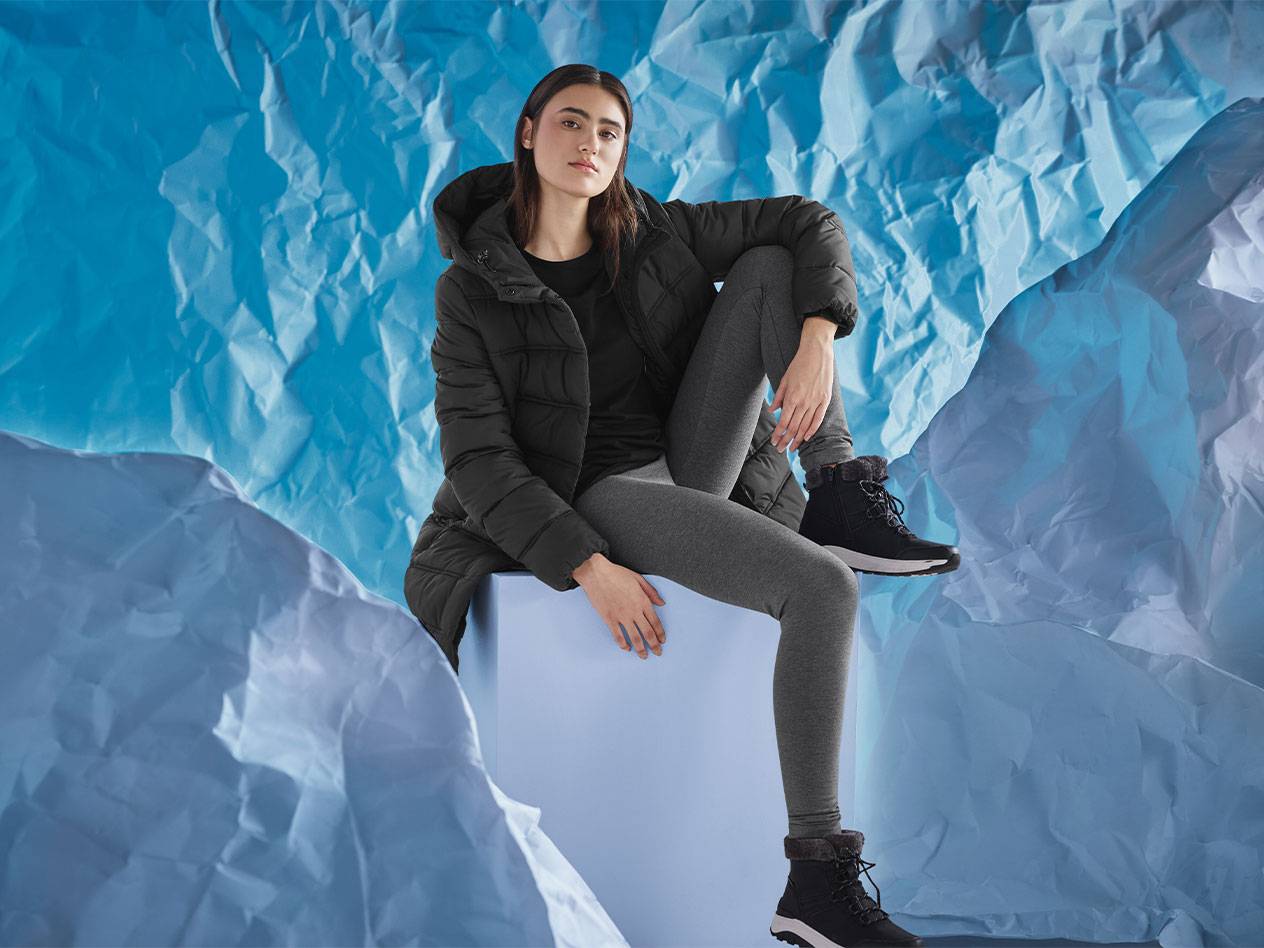 Woman in black puffer jacket, grey leggings, and black winter boots sitting on a blue box.