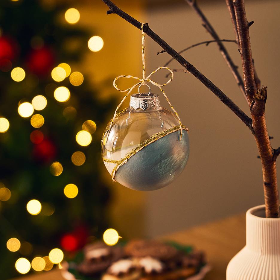 Transparent and blue Christmas bauble with gold glitter, hanging from a branch with Christmas lights in background.
