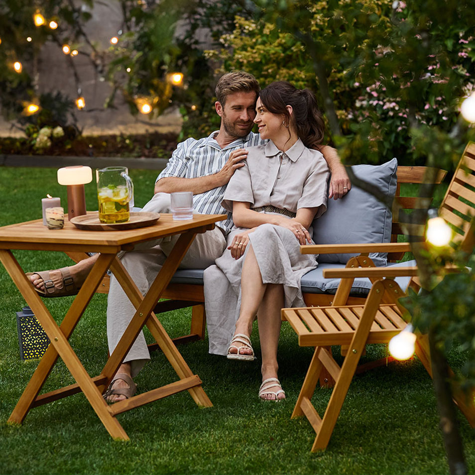 Couple relaxing in a garden on wooden outdoor furniture with lighting.