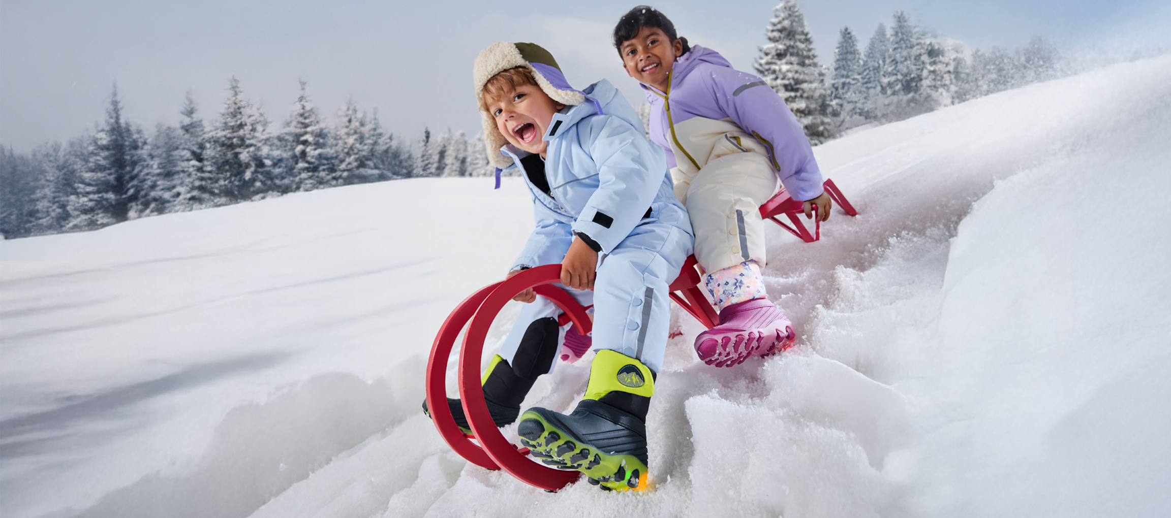 Two children in snowsuits and winter boots, sledding down a snowy slope.