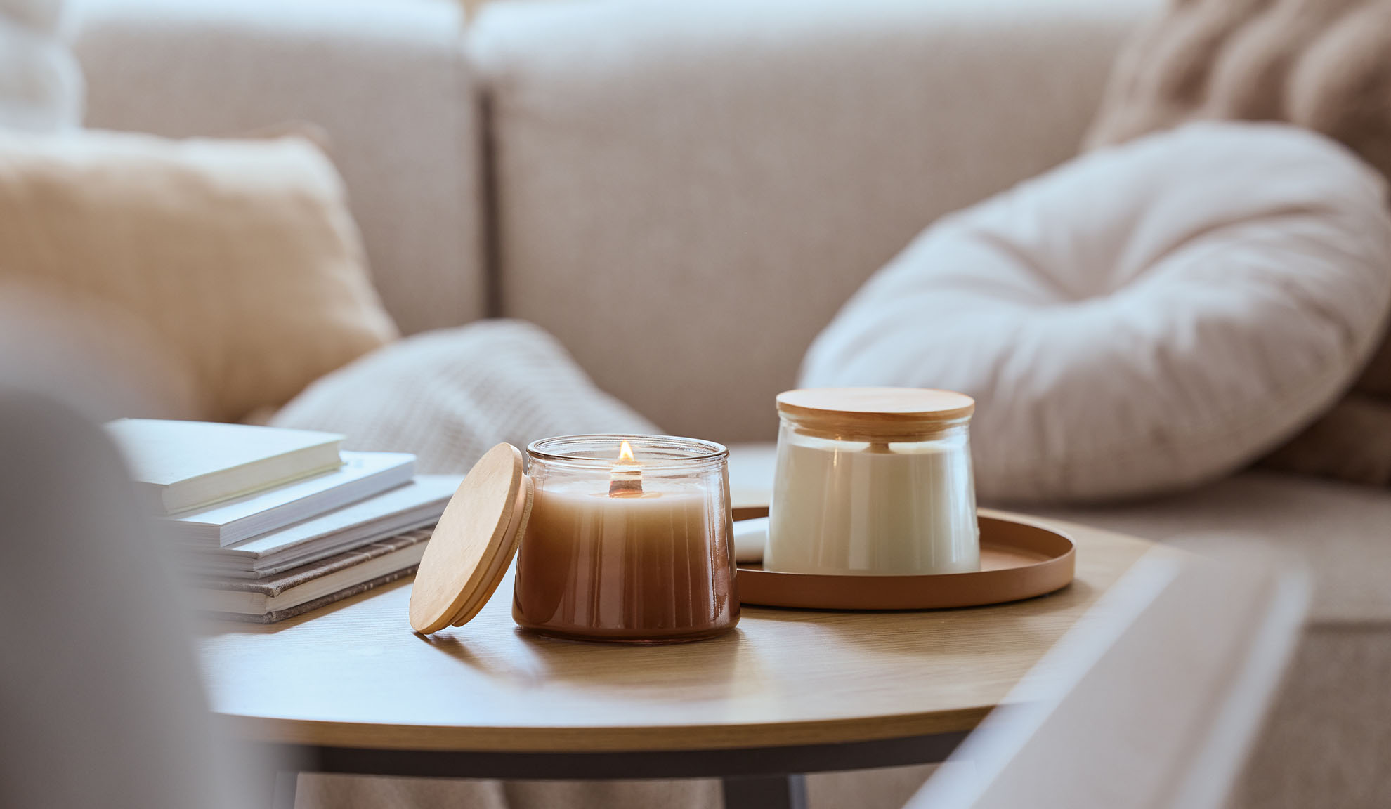 Two scented candles on a wooden table, one lit, with a cozy sofa in the background.