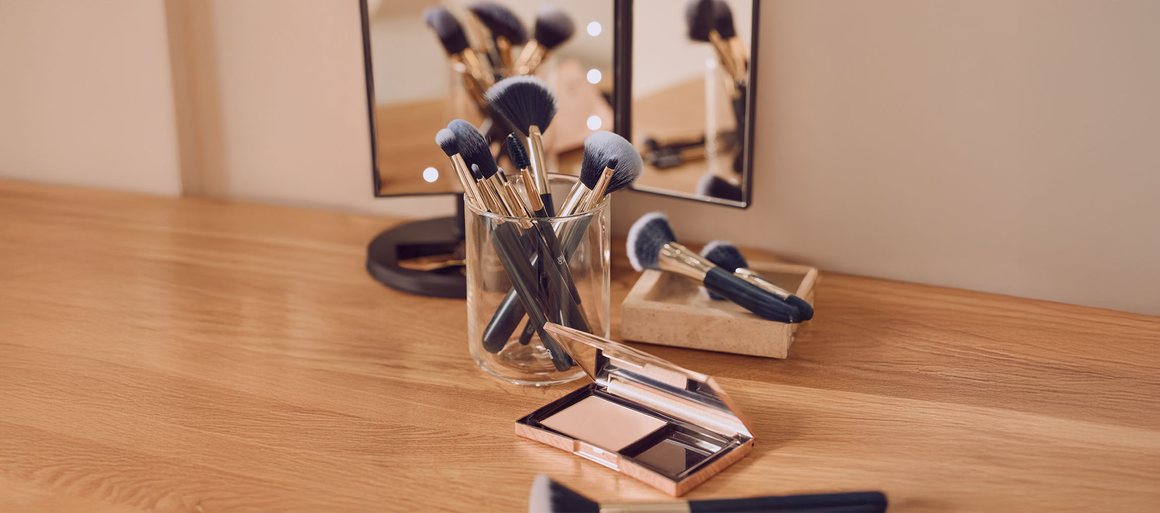 Makeup brushes in a glass holder and compact powder on a wooden vanity table.