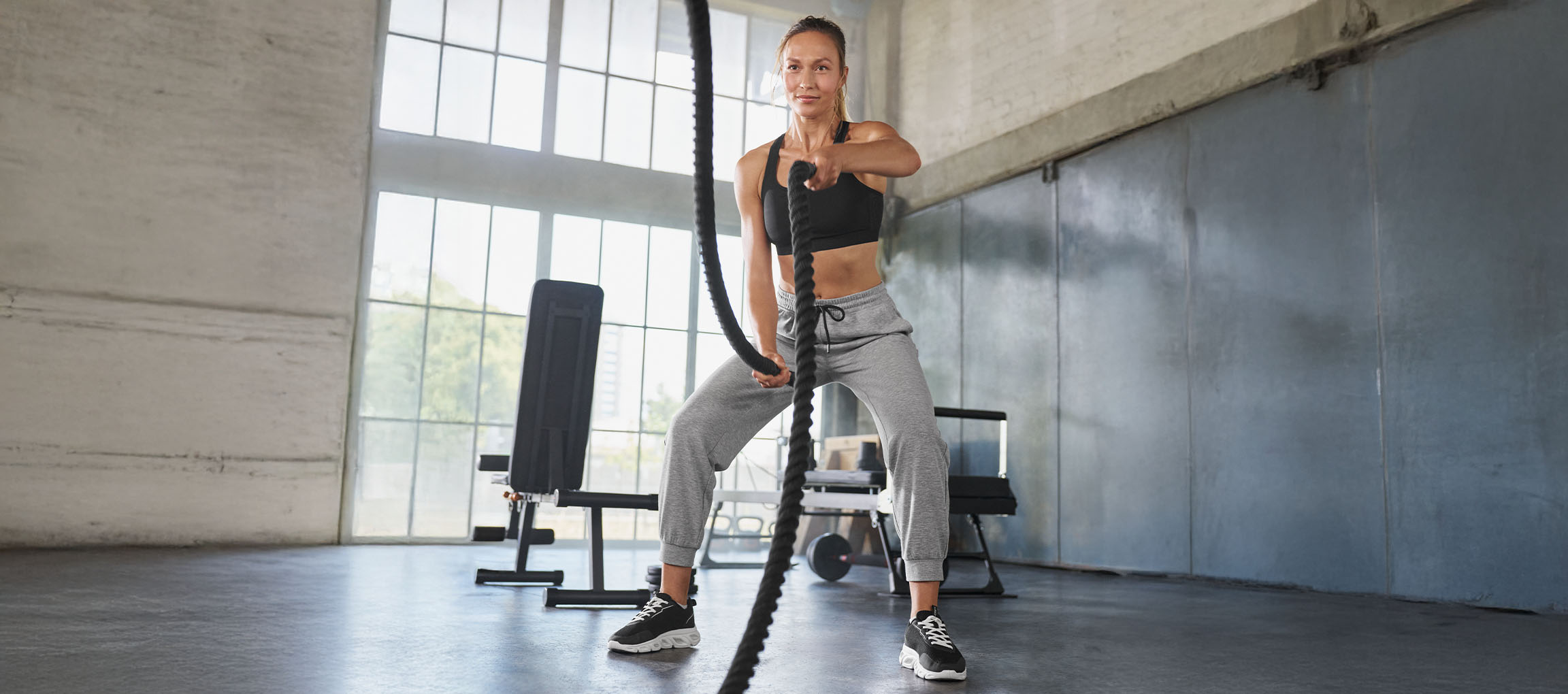 Woman in black sports bra and grey sweatpants exercising with battle ropes