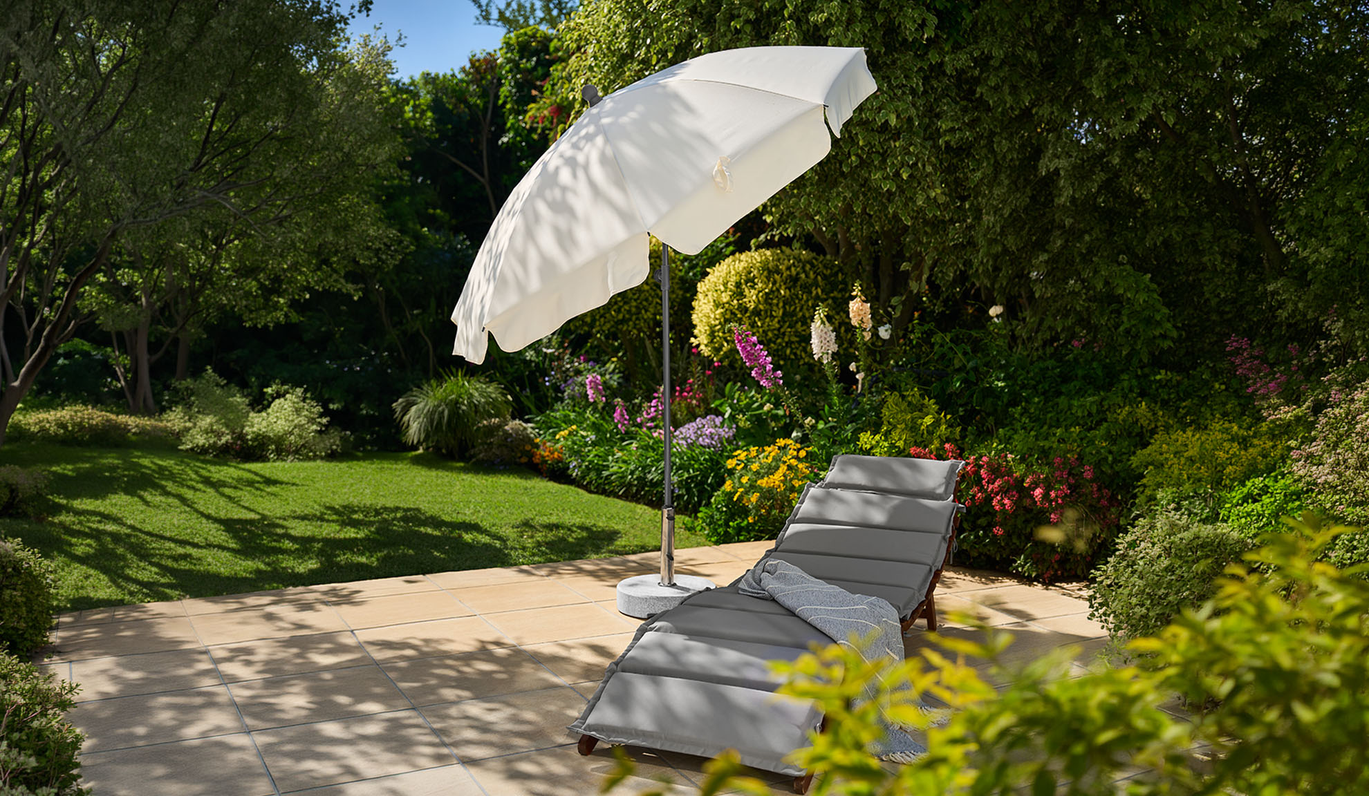 Grey sun lounger with white parasol on a sunny patio with a lush garden.