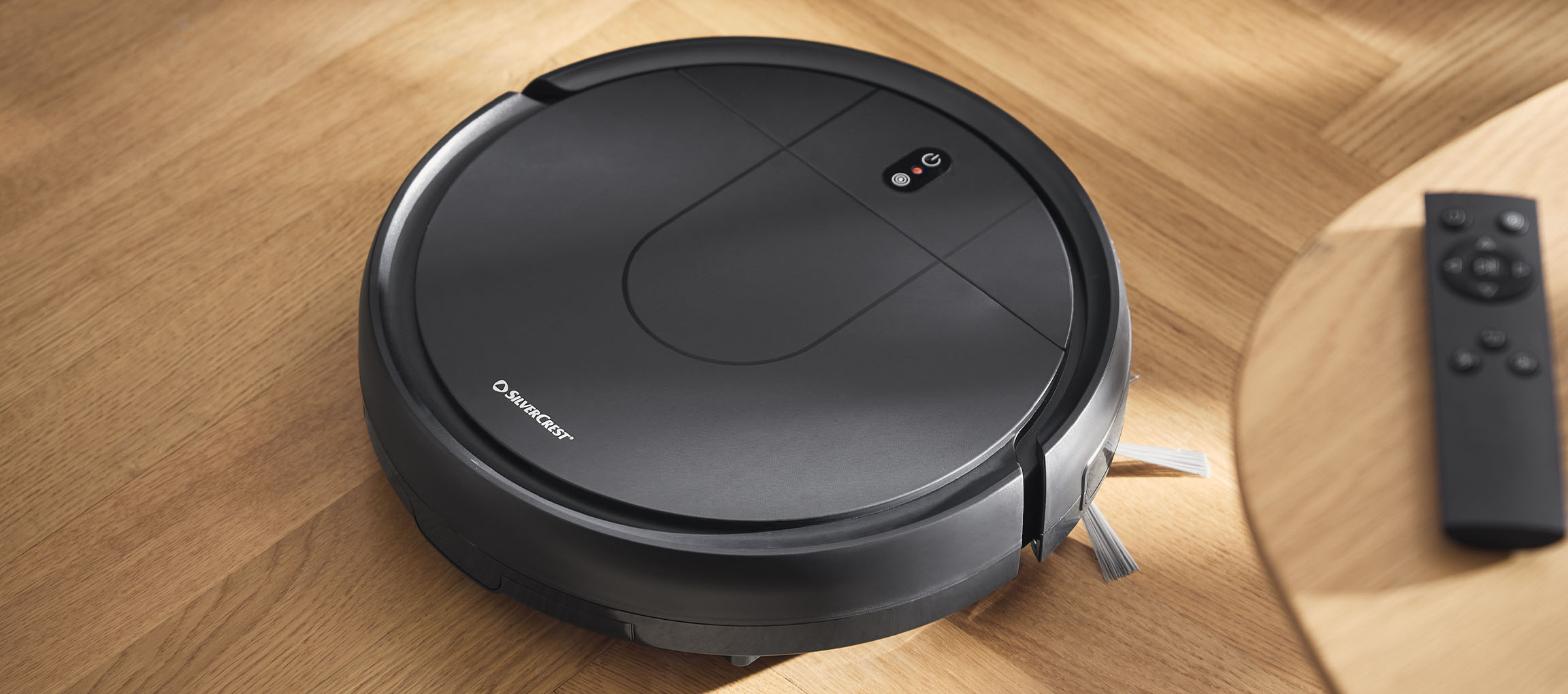 Black robotic vacuum cleaner on a wooden floor with a remote control