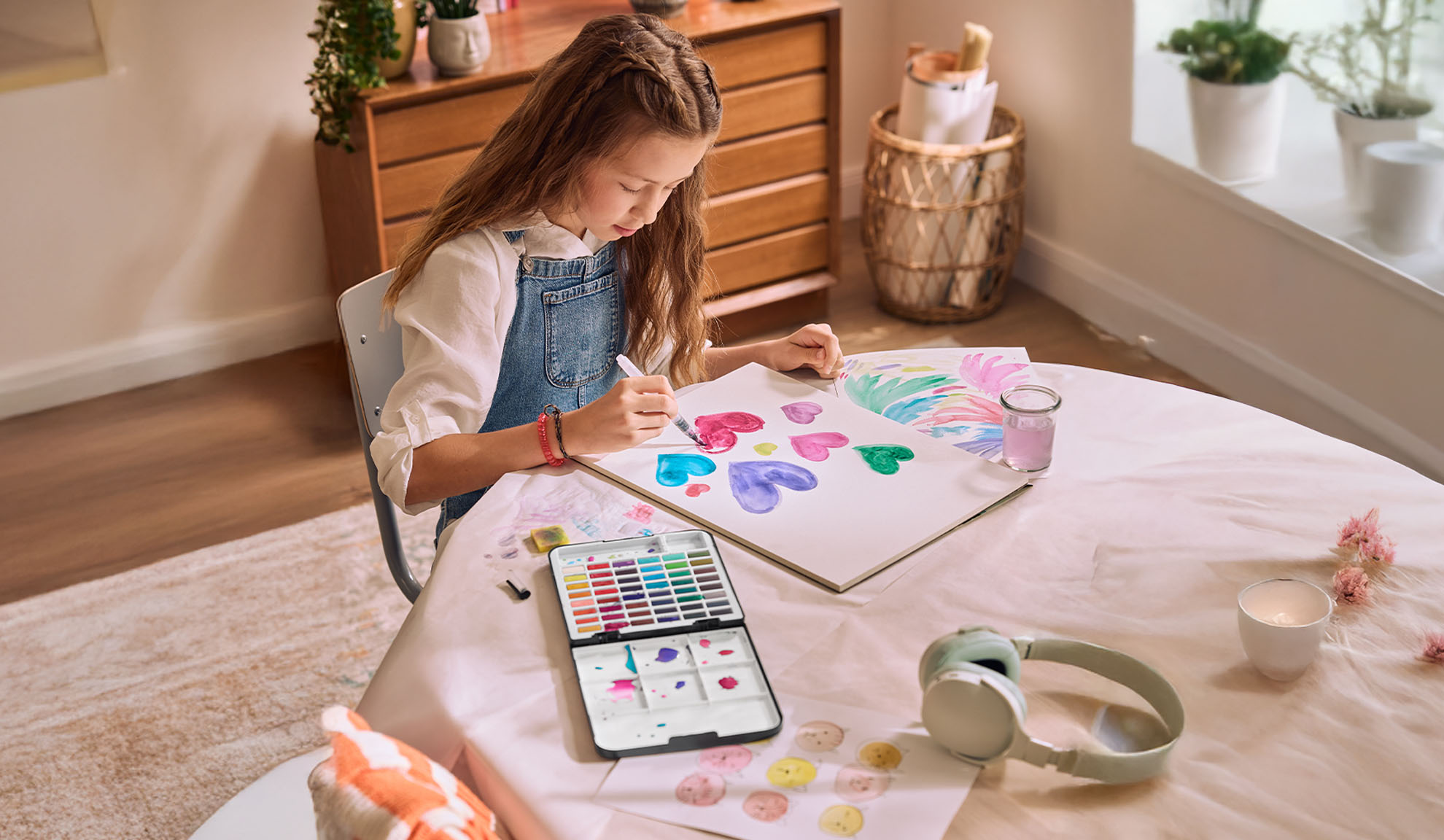 A girl painting hearts with watercolors at a table, with a watercolor set and headphones nearby.