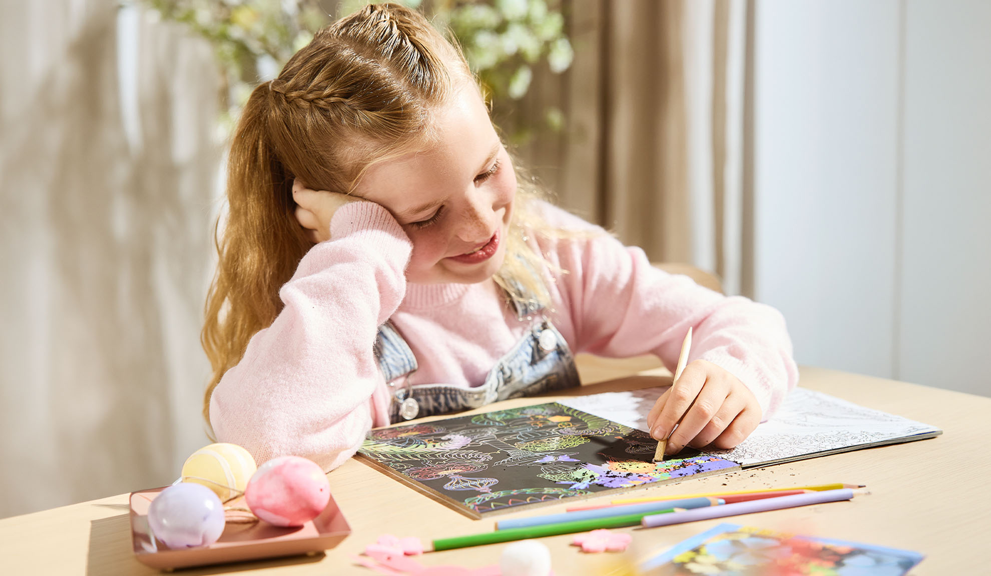 Girl scratching a colourful scratch art picture, with bath bombs and pencils on the table.