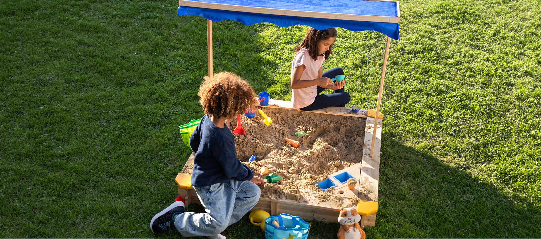 Two children playing in a wooden sandbox with a blue canopy, surrounded by green grass.