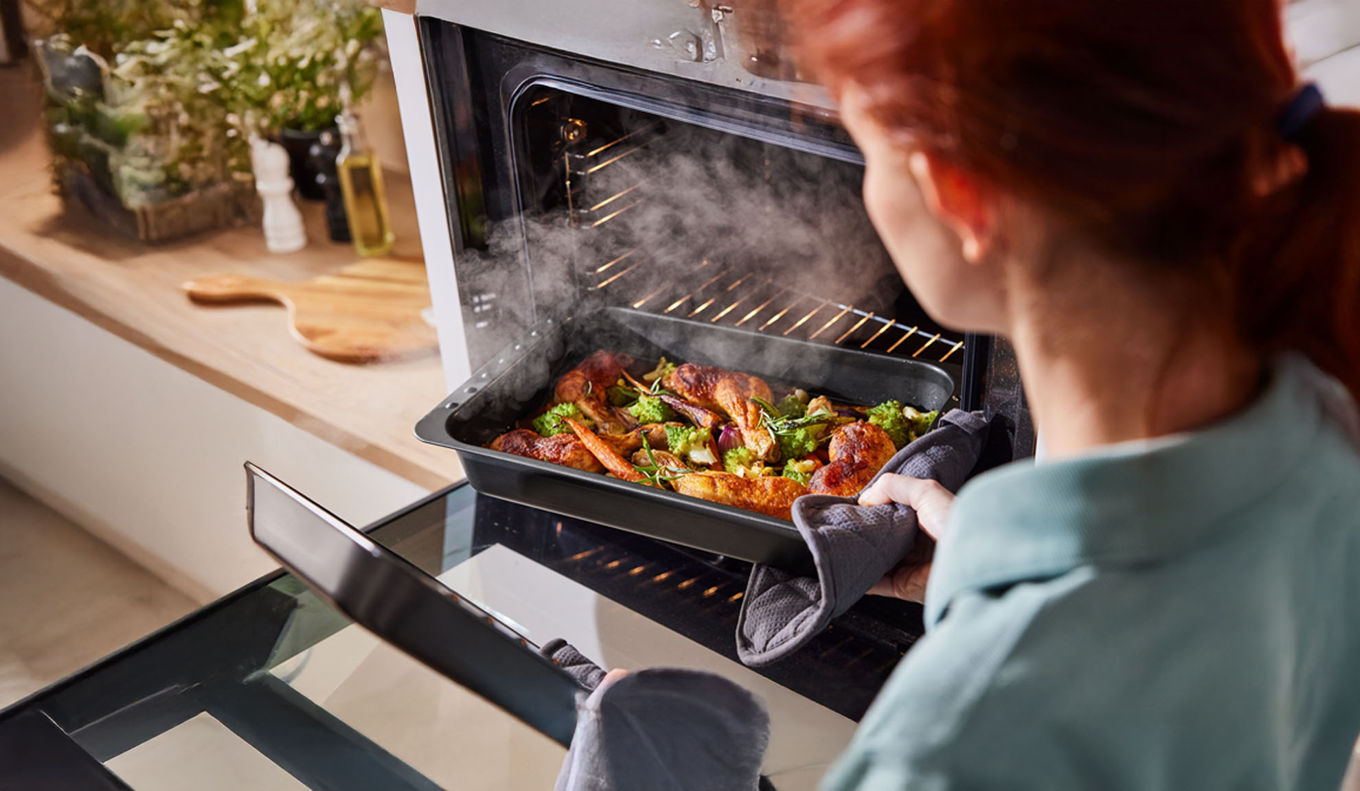 Woman removing a steaming tray of chicken and vegetables from an oven.