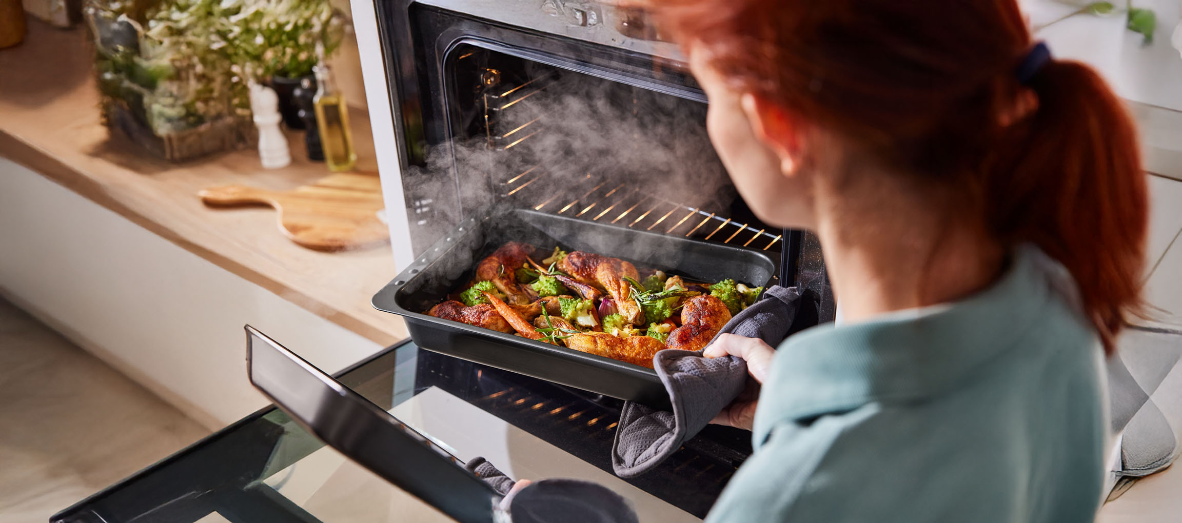 Woman removing a baking tray of chicken and vegetables from a steaming oven.