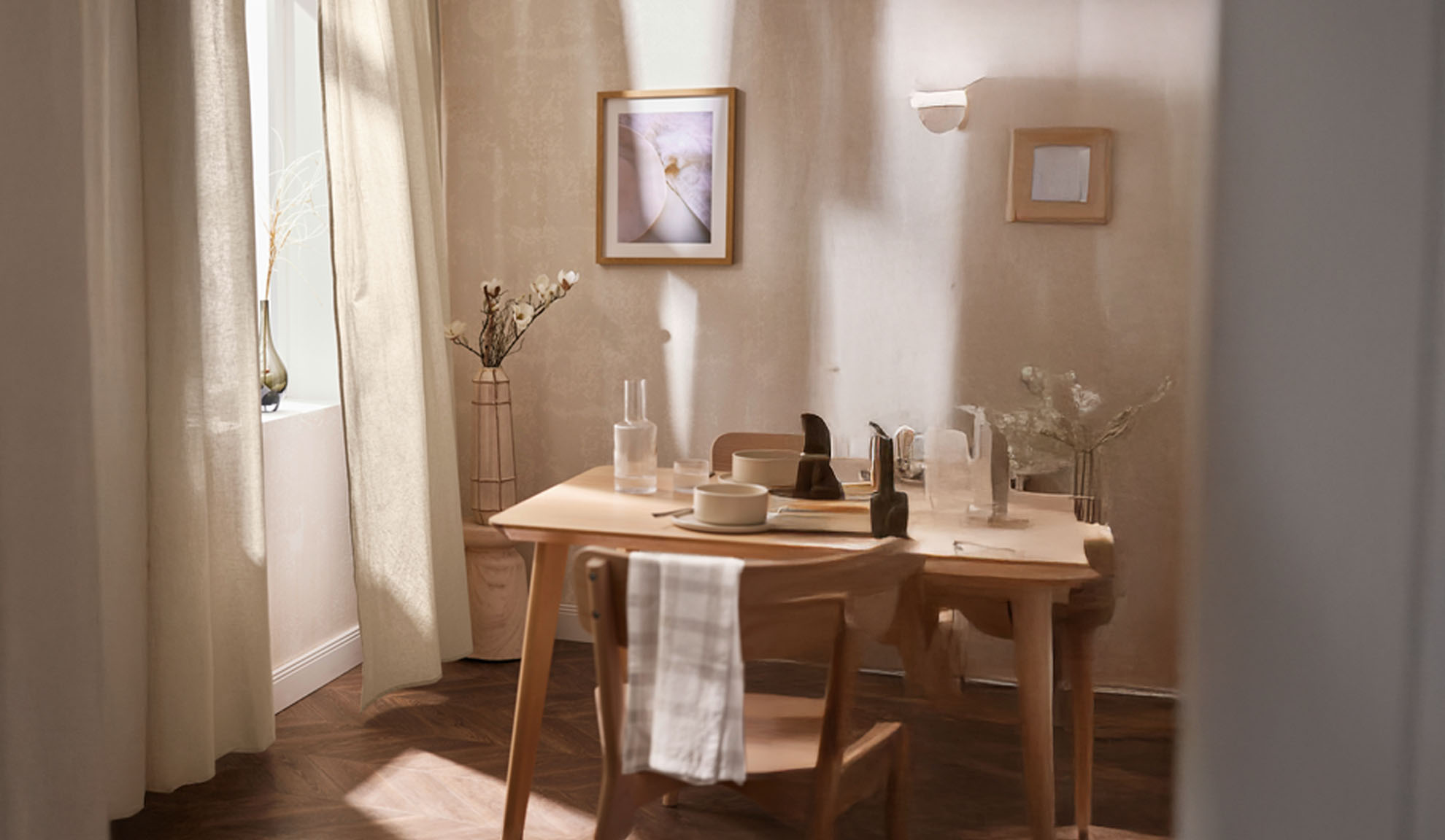 A dining room with a wooden table set for two, featuring light-colored decor and natural light.