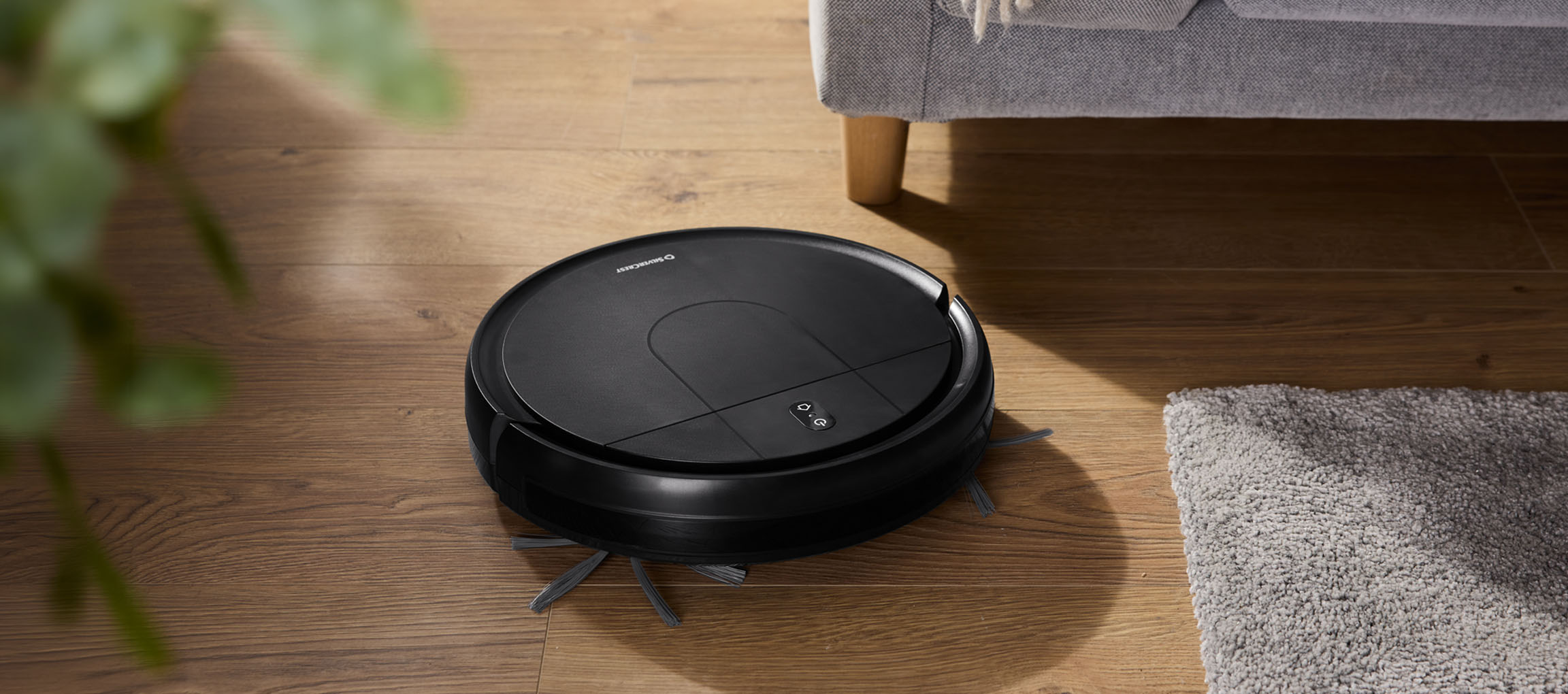 Black robotic vacuum cleaner on a wooden floor next to a sofa and a rug.
