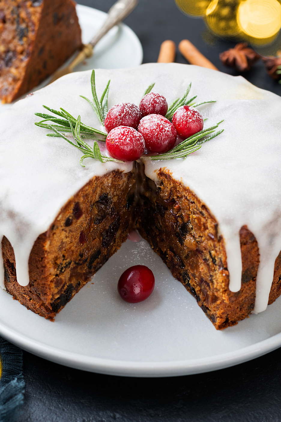 Christmas fruitcake with white icing, cranberries, and rosemary, with a slice removed.