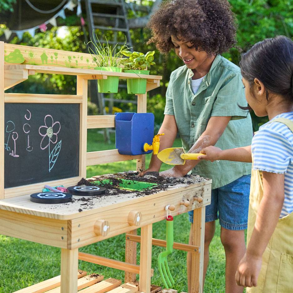 Two children playing with a wooden mud kitchen, featuring a chalkboard and potted plants.