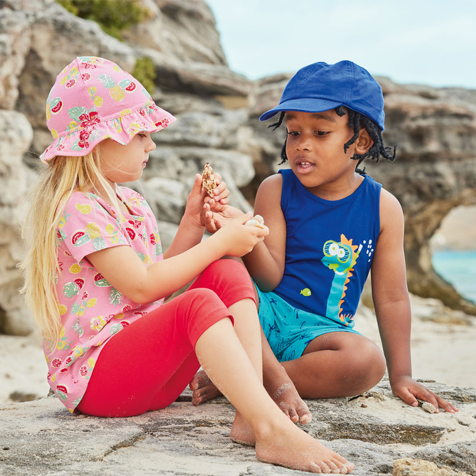 Two children on a beach, one in a pink sun hat and fruit-patterned top, the other in a blue cap and dinosaur tank top, holding seashells.