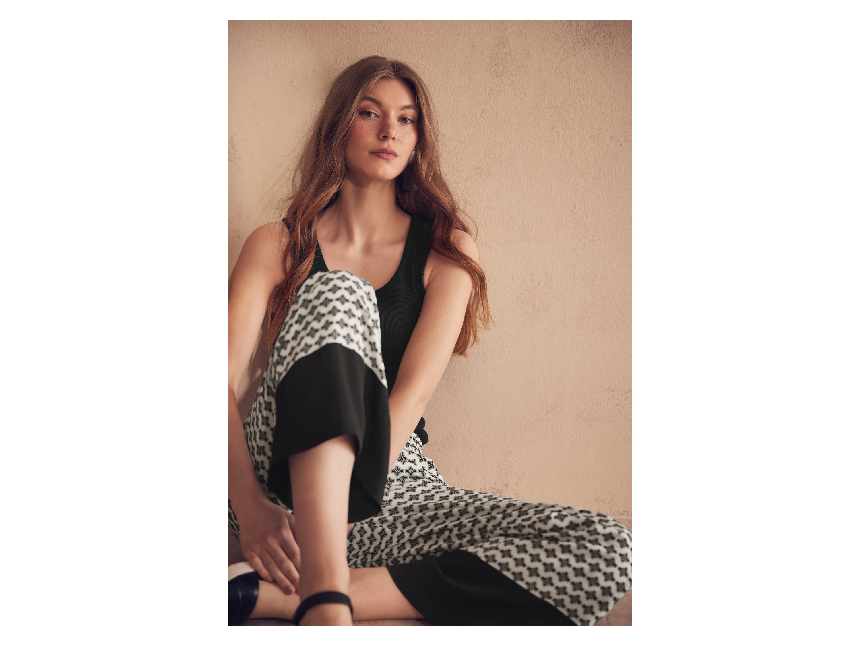 Woman in black tank top and patterned pants sits against a beige wall.