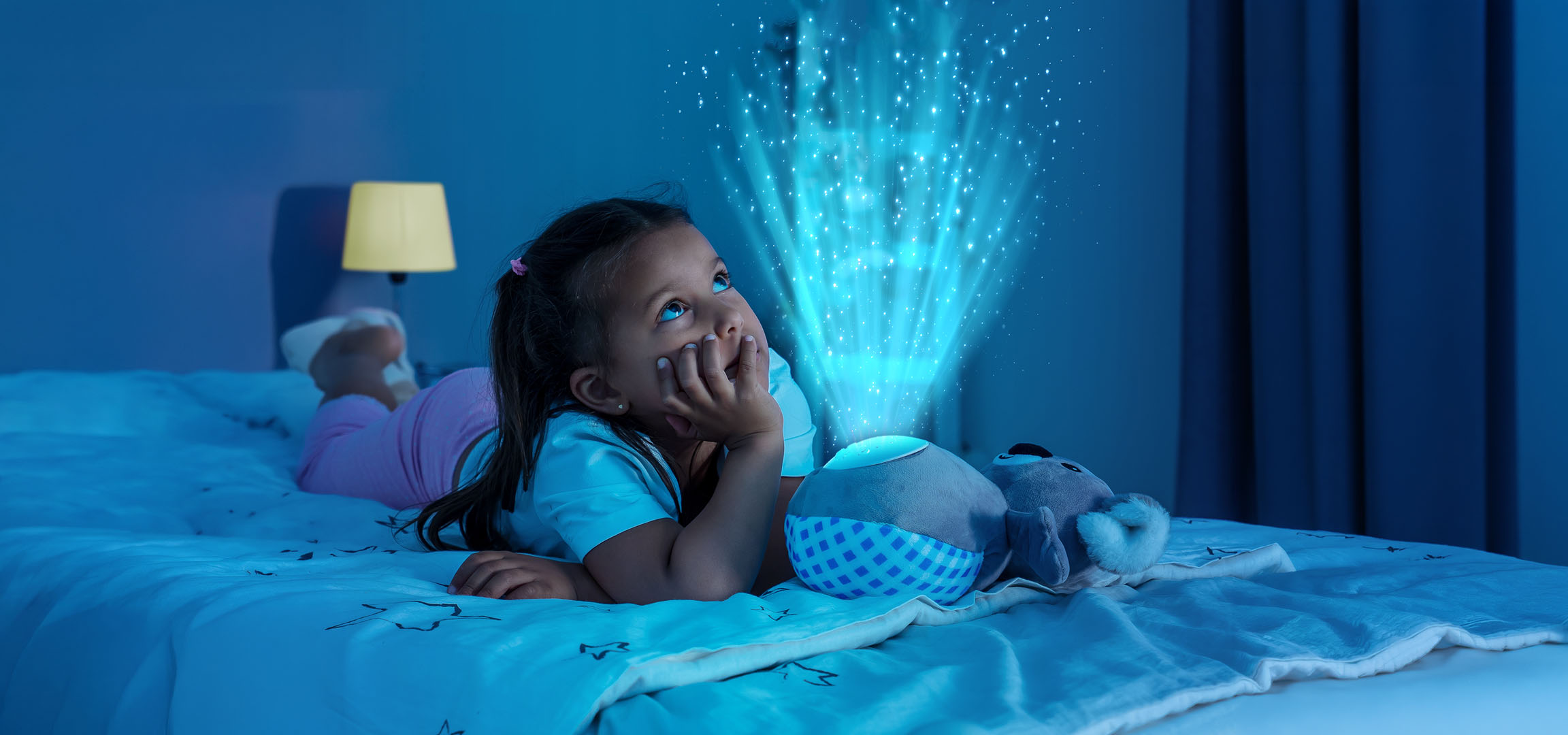Girl lying in bed, looking at a star projector casting lights on the wall.