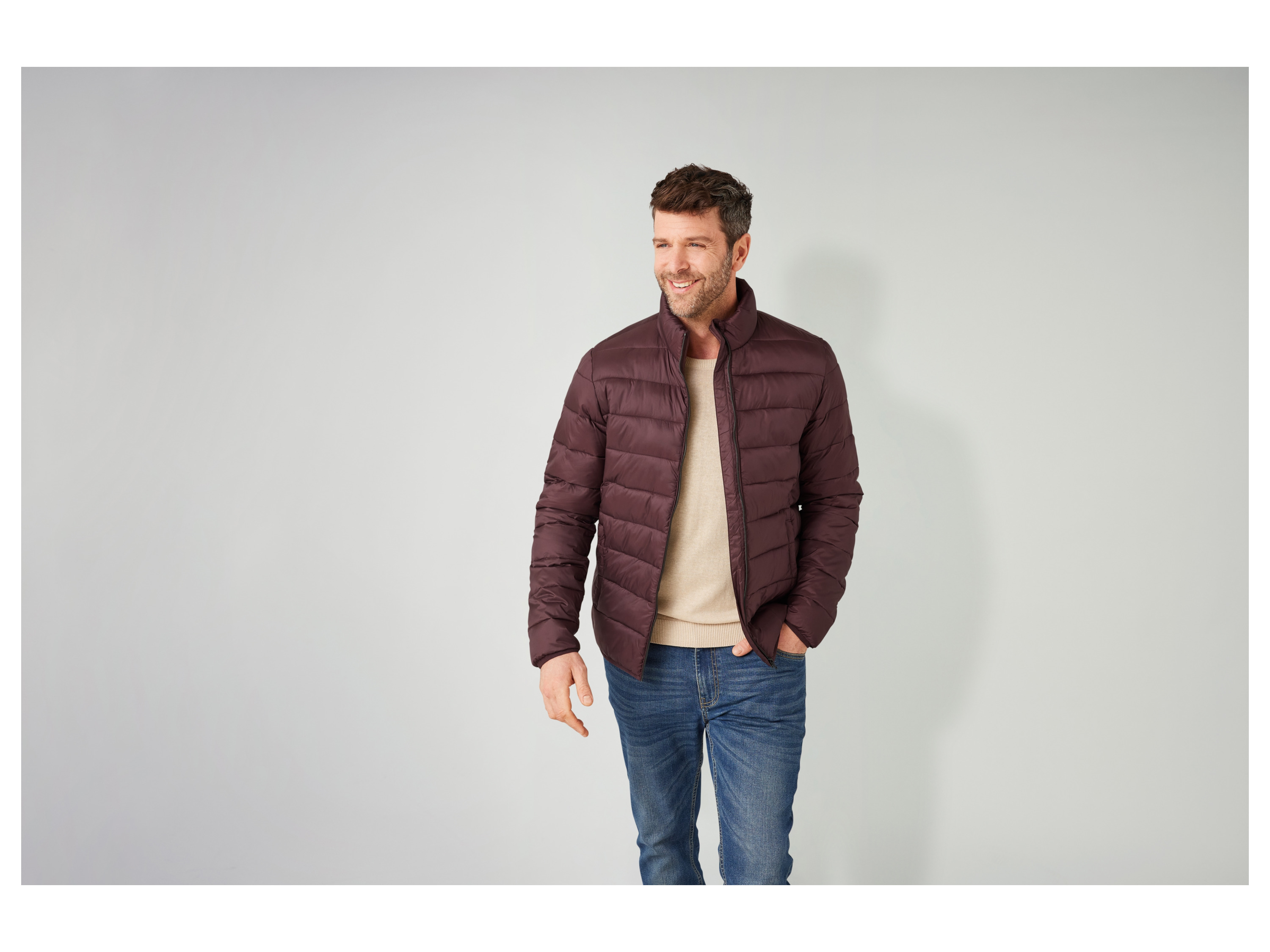 A man smiles while wearing a burgundy puffer jacket and blue jeans.